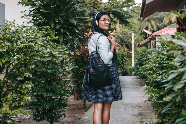 Young stylish woman walking, listening to music on headphones, smiling, happy, holding backpack, summer vacation