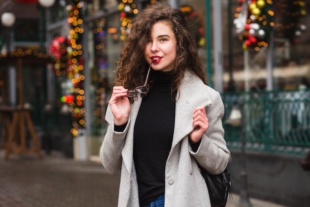 Young stylish woman walking in street in autumn grey coat, curly hair, cold weather, fashion trend casual style