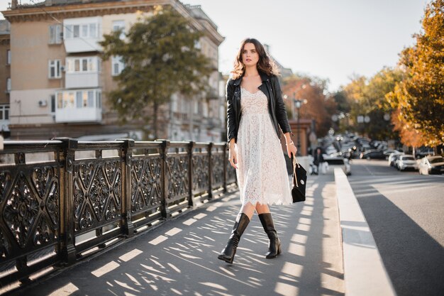 Young stylish woman walking in street in fashionable outfit, holding purse, wearing black leather jacket and white lace dress, spring autumn style