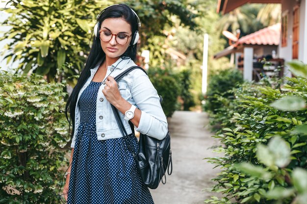 Young stylish woman walking with smartphone, listening to music on headphones, summer vacation