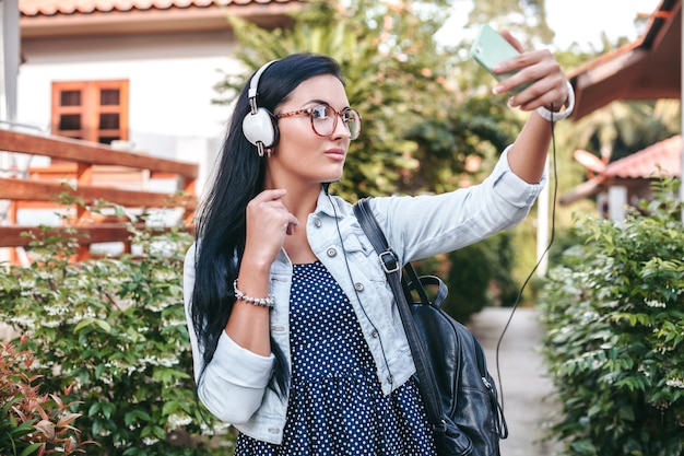 Young stylish woman walking with smartphone, listening to music on headphones, taking photo, vintage denim style, summer vacation