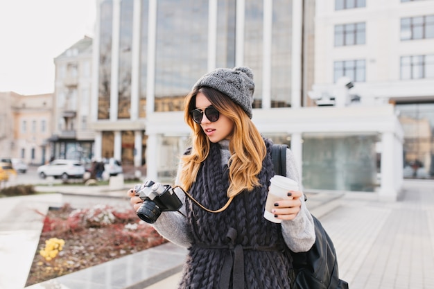 Young stylish woman in warm woollen sweater, modern sunglasses and knitted hat walking with coffee to go in city centre. Travelling with backpack, tourist with camera, cheerful mood.
