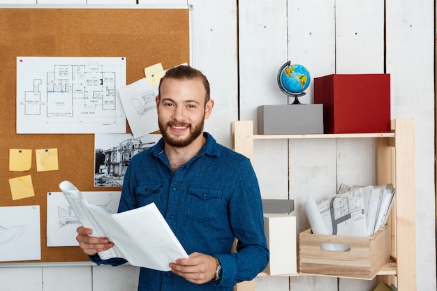 Young successful architector smiling, holding drawings, standing in office wall