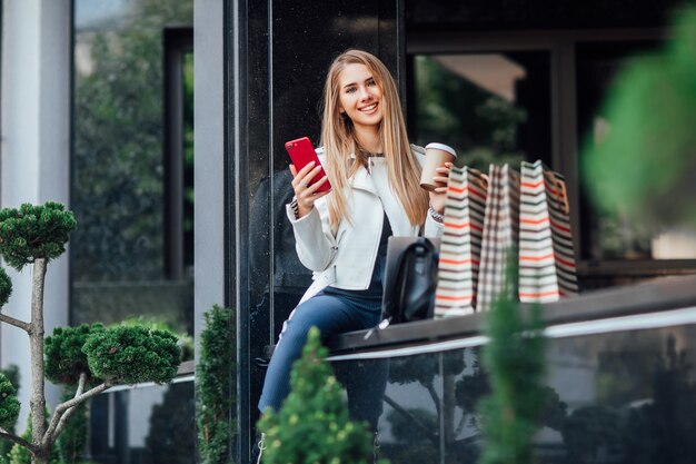 젊고 성공적인 금발의 유행 여성은 커피와 전화 한 잔을 들고 쇼핑 시간을 보낸 후 가게 근처에 앉아 있습니다.
