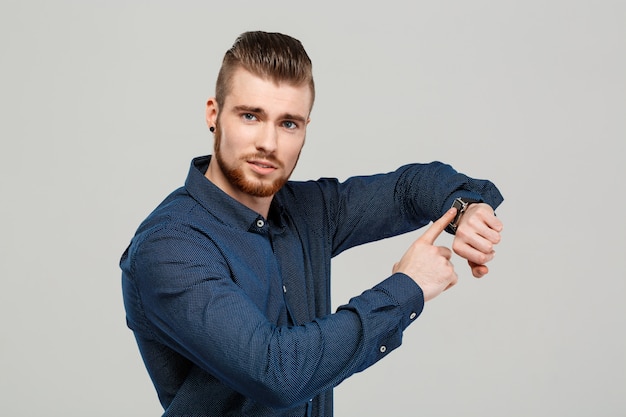 Free photo young successful businessman pointing at watch over grey wall.