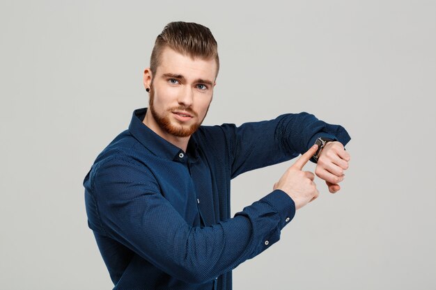 Young successful businessman pointing at watch over grey wall.