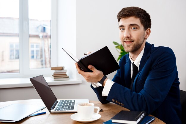 Young successful businessman sitting at workplace, office background.