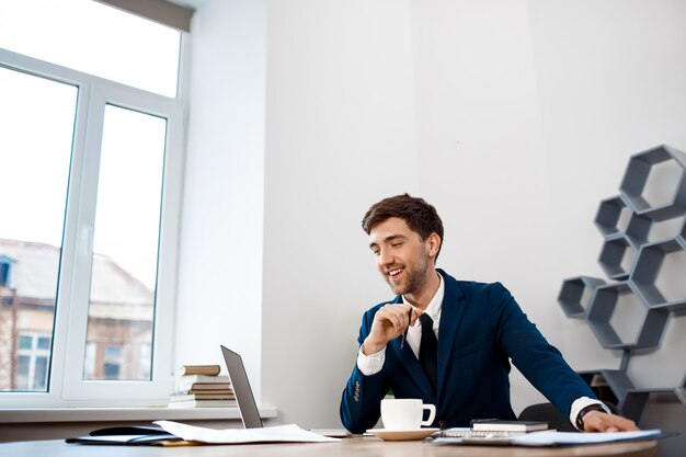 Young successful businessman sitting at workplace, office background.