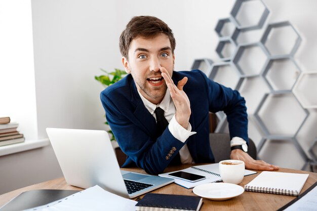 Young successful businessman sitting at workplace, office background.