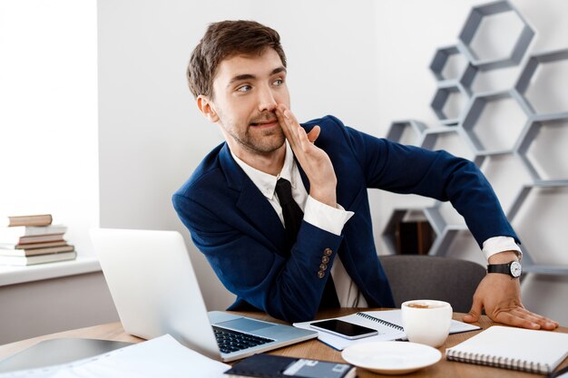 Young successful businessman sitting at workplace, office background.