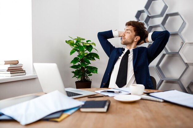 Young successful businessman sitting at workplace, office background.