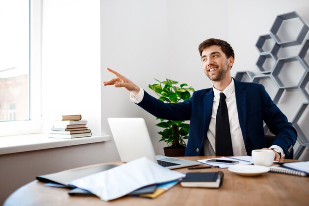 Young successful businessman sitting at workplace, office background.