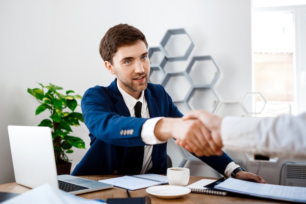 Young successful businessman sitting at workplace, office background.