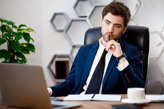 Young successful businessman sitting at workplace, office background.