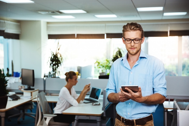 Young successful businessman smiling, looking at tablet, over office