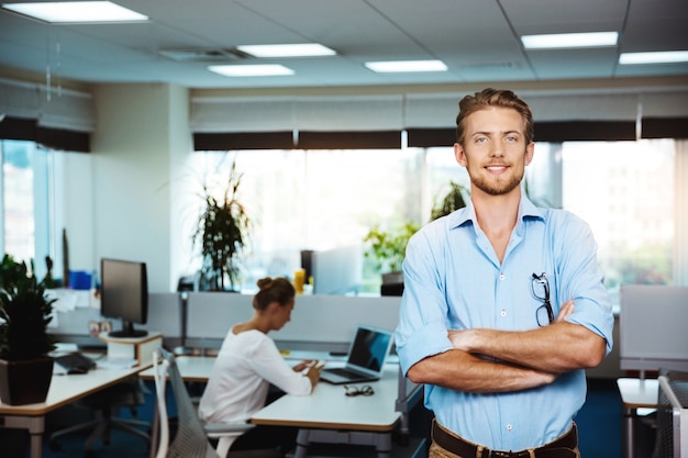 Free photo young successful businessman smiling, posing with crossed arms, over office