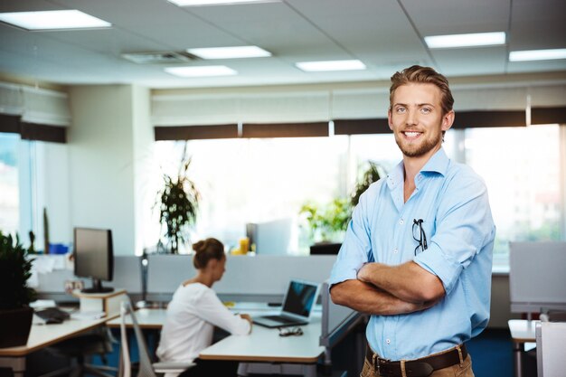 Young successful businessman smiling, posing with crossed arms, over office