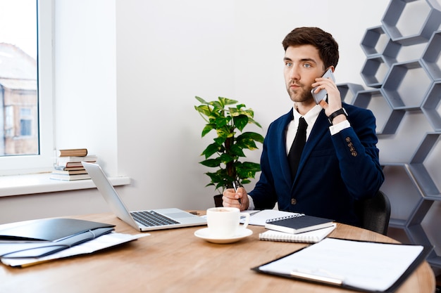 Giovane riuscito uomo d'affari che parla sul telefono, fondo dell'ufficio.