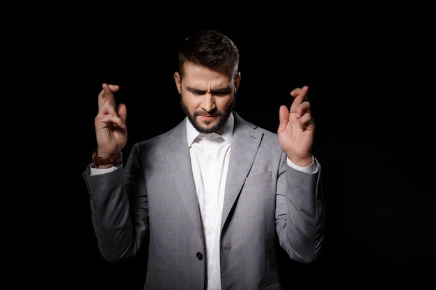 Young successful businessman in suit praying with crossed fingers isolated on black