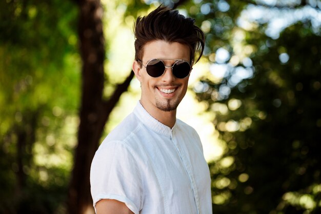 Young successful businessman in sunglasses smiling, walking around sunny city.