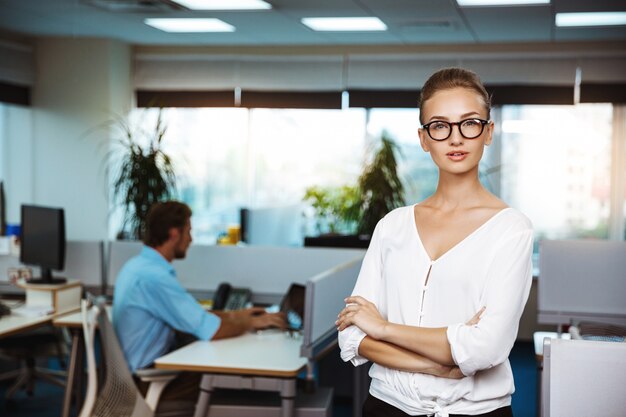 Young successful businesswoman smiling, posing with crossed arms, over office