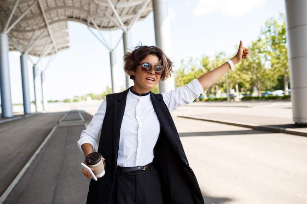 Young successful businesswoman in sunglasses catching car over business centre.