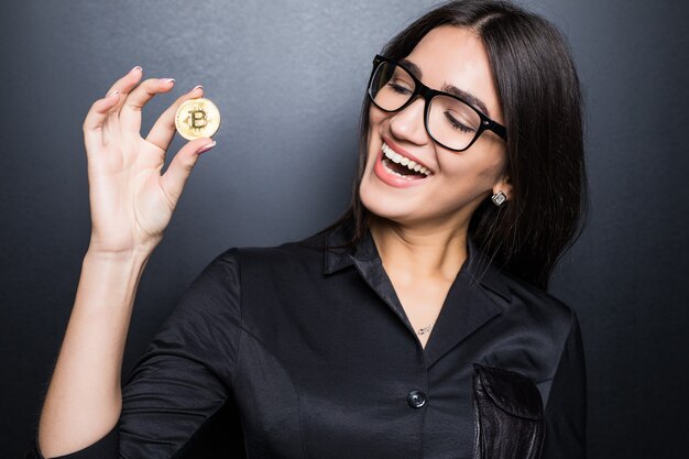 Young successful confident woman with glasses holds a gold bitcoin in her hand isolated on black wall