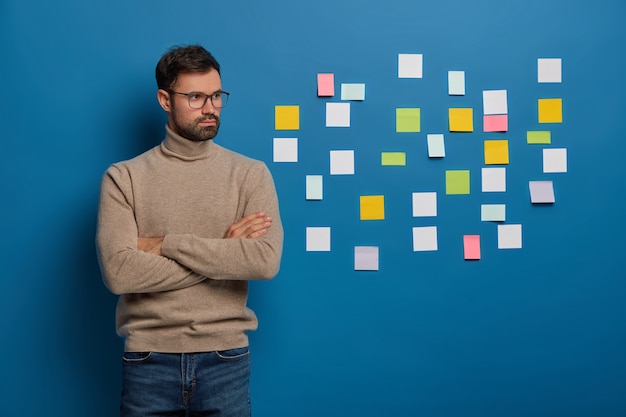Free photo young successful entrepreneur stands in thoughtful pose, brainstorms ideas for developing startup project, stands with crossed hands against blue background, sticky notes stuck on blue wall behind