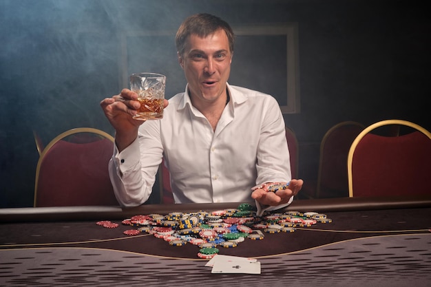 Young successful guy in a white shirt is playing poker sitting at the table at casino in smoke. He rejoices in winning showing his chips and holding a glass of whiskey in his hand. Gambling for money.