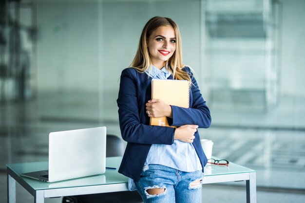 Young successful woman in casual clothes holding notebook work standing near white desk with laptop in office. Achievement business career concept.