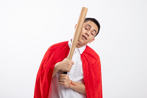 Free photo young superhero boy in red cape beating himself in face with baseball bat with closed eyes isolated on white background with copy space