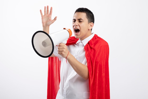 Young superhero boy in red cape standing in profile view looking straight shouting in loud speaker raising hand up isolated on white background with copy space