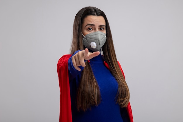 白い背景で隔離のジェスチャーを示す医療マスクを身に着けている若いスーパーヒーローの女の子