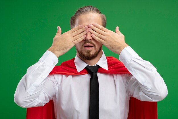 Young superhero guy covered eyes with hands isolated on green