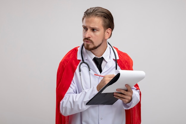 Young superhero guy looking at side wearing medical robe with stethoscope holding pen