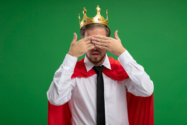 Young superhero guy wearing crown and tie covered eyes with hands isolated on green