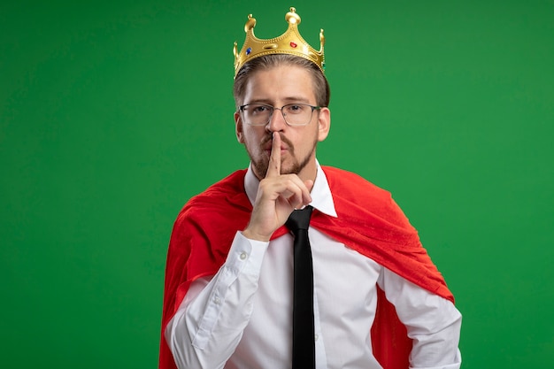 Free photo young superhero guy wearing crown and tie showing silence gesture isolated on green