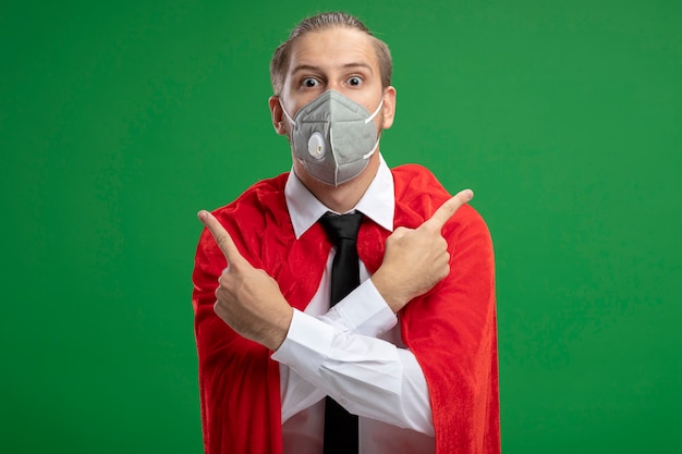 Free photo young superhero guy wearing medical mask and tie points at differents sides isolated on green