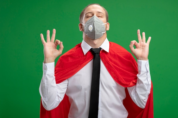 Ragazzo giovane supereroe che indossa maschera medica e cravatta con gli occhi chiusi facendo meditazione isolato su sfondo verde