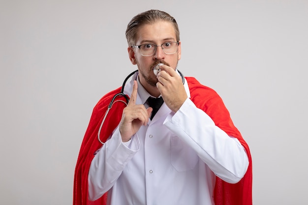 Young superhero guy wearing medical robe with stethoscope and glasses putting stethoscope on mouth and showing silence gesture isolated on white background