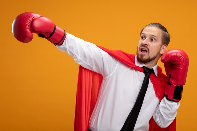 Young superhero guy wearing tie and boxing gloves looking at side holding out hand at side isolated on orange