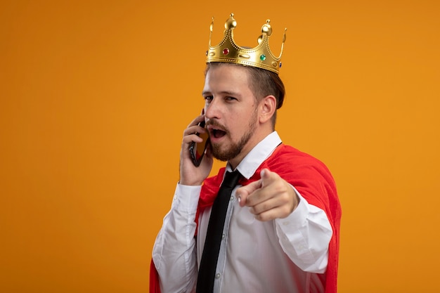 Ragazzo giovane supereroe indossa cravatta e corona che guarda l'obbiettivo parla sul telefono che mostra il gesto isolato su sfondo arancione
