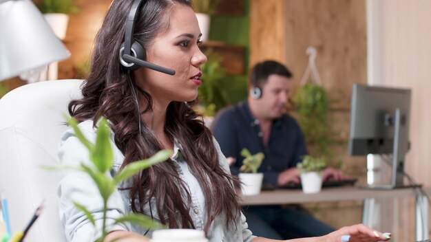 Young support customer woman talking into headset microphone with call center customer while working in startup company office. Caucasian female consultant providing telemarketing service