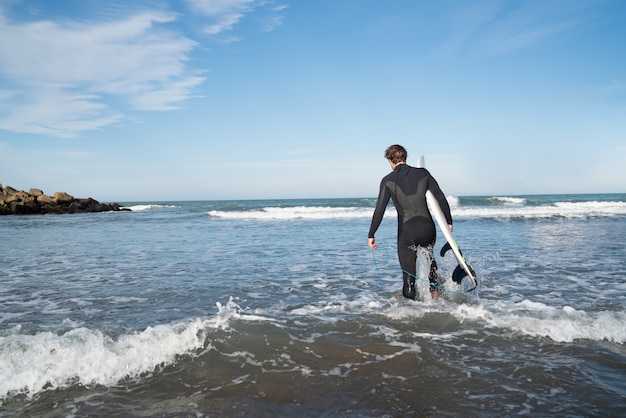 검은 서핑 정장을 입고 자신의 서핑 보드와 함께 물에 들어가는 젊은 서퍼. 스포츠 및 수상 스포츠 개념.