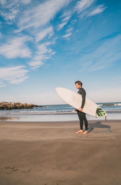 Young surfer standing in the ocean with his surfboard in a black surfing suit. Sport and water sport concept.