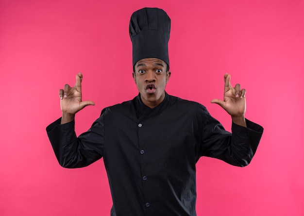 Free photo young surprised afro-american cook in chef uniform crosses fingers of both hands isolated on pink wall