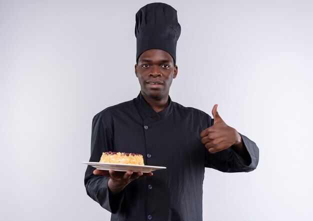 Young surprised afro-american cook in chef uniform holds cake on plate and thumbs up on white  with copy space