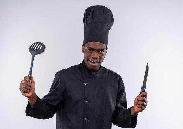 Il giovane cuoco afroamericano sorpreso in uniforme del cuoco unico tiene la spatola del nd del coltello su bianco con lo spazio della copia