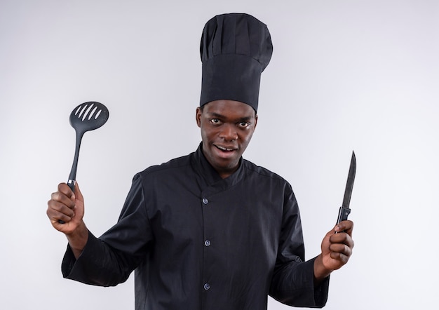 Young surprised afro-american cook in chef uniform holds knife nd spatula on white  with copy space