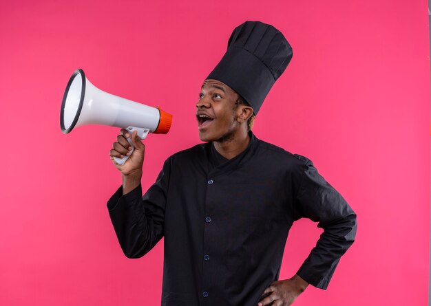 Young surprised afro-american cook in chef uniform holds loud speaker isolated on pink wall 